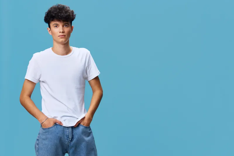 Young man in casual white t-shirt and blue jeans standing with hands in pockets against blue background, looking confident and relaxed, lifestyle portrait.