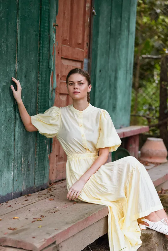 A woman in a pale yellow dress sits gracefully on a porch leaning against a green wall, creating a calm outdoor portrait with thoughtful serene poise