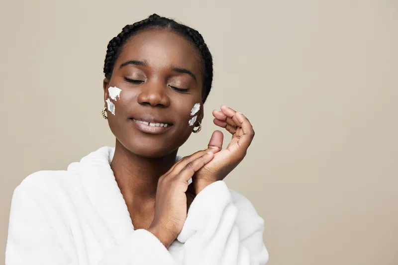 Relaxed African woman applying skincare cream in a cozy robe, enjoying self care routine with a serene expression and neutral background