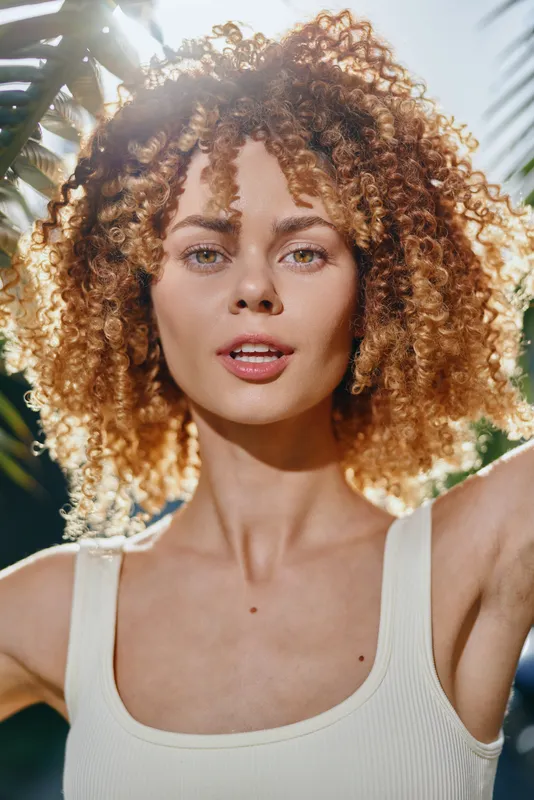 Young woman with curly hair posing outdoors with natural sunlight, wearing a white tank top and surrounded by green leaves in a summer setting.