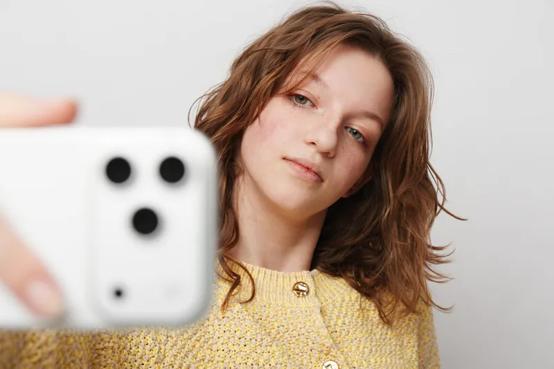 Young woman with wavy brown hair wearing yellow sweater takes selfie with smartphone on white background. Concept of casual lifestyle, technology, and self-portrait photography.