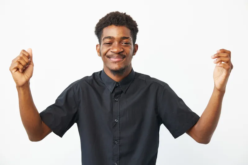 Happy African American man celebrating with raised arms, dressed in a black shirt, expressing joy and positivity against a clean white background.