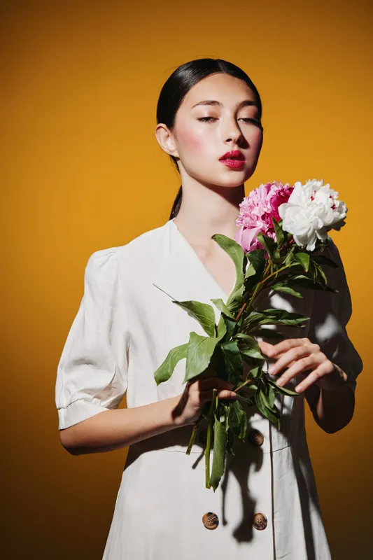 Young woman with red lipstick holding pink and white peonies wearing a white dress against yellow background. Concept of beauty, fashion, and creative floral style.