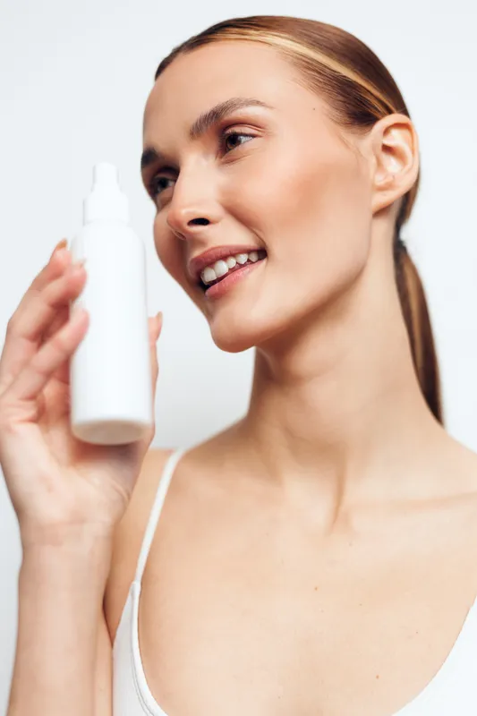 Young woman with clear skin holds white cosmetic bottle smiling gently isolated on white background skincare concept beauty product for face care and daily routine