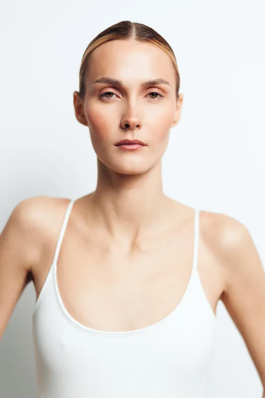 A young woman stands against a plain light backdrop wearing a white tank top, exuding calm confidence and modern minimalism in a clean studio portrait.