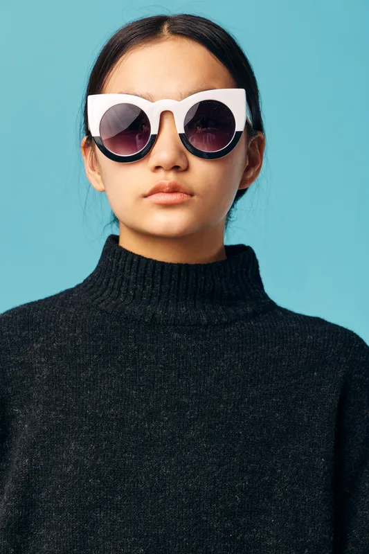 Young woman in oversized white sunglasses and black turtleneck sweater posing against blue background with serious expression and natural makeup closeup portrait in studio.