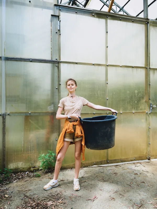 Woman in greenhouse holding a large black bucket, young gardener in casual clothes with jacket tied at waist, plants and soil visible on earthy ground under translucent panels.