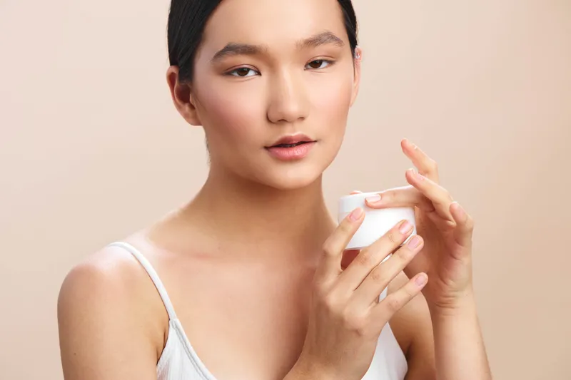 asian woman overlooks a softly lit space as she applies skincare, a gentle smile and calm expression hinting at a thoughtful selfcare moment in a private, intimate setting.