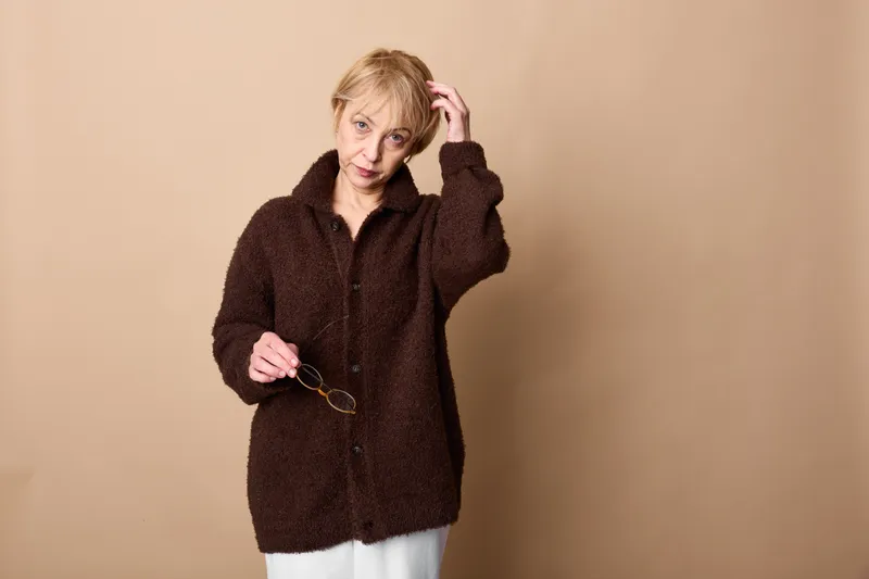 Woman with glasses and coat portrait of blonde adult in studio, beige background, she adjusts eyewear with calm smile, soft light, winter fashion, comfort and warmth