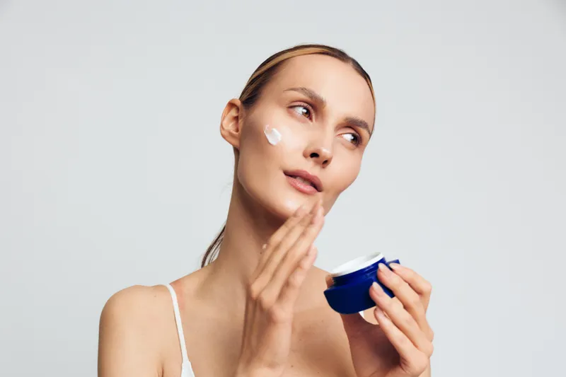 Young woman applying face cream on clean skin, skincare routine, natural beauty, soft lighting, white background, focused expression, healthy complexion