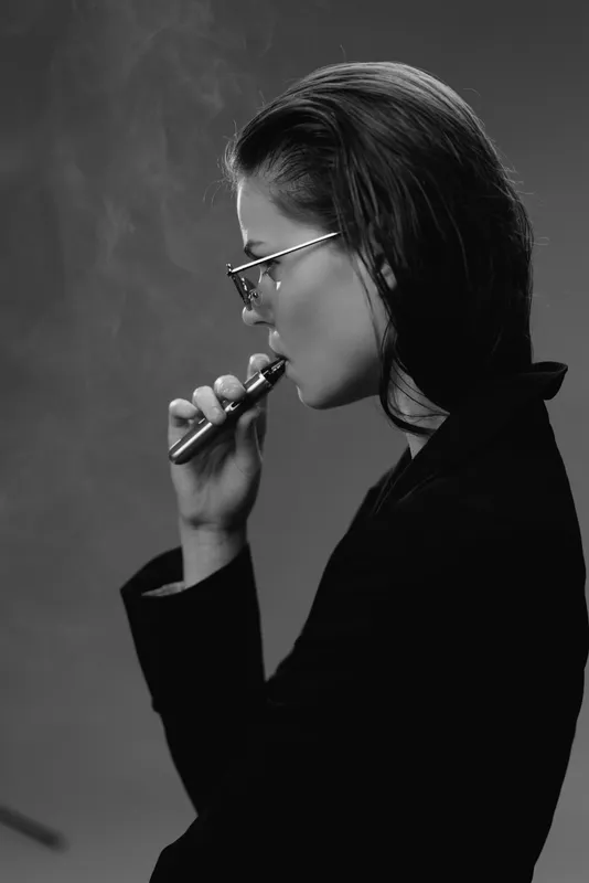 Black and white image of a young woman vaping with glasses and dark hair, wearing a black outfit in a smoky atmosphere.