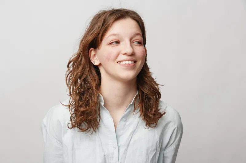 Young woman with curly brown hair wearing a white shirt smiles gently looking to the side. Studio portrait on a light grey background with natural soft lighting and casual style.