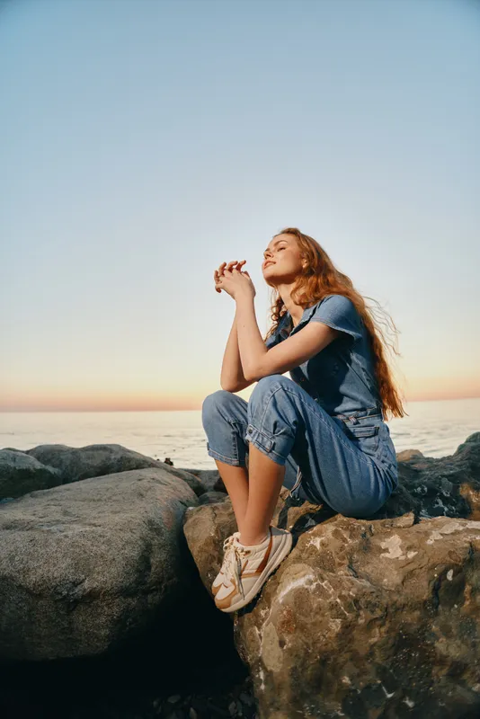 Lifestyle woman in modern boho-western denim street style sitting on rocks by the sea during golden hour with relaxed expression and film color tones