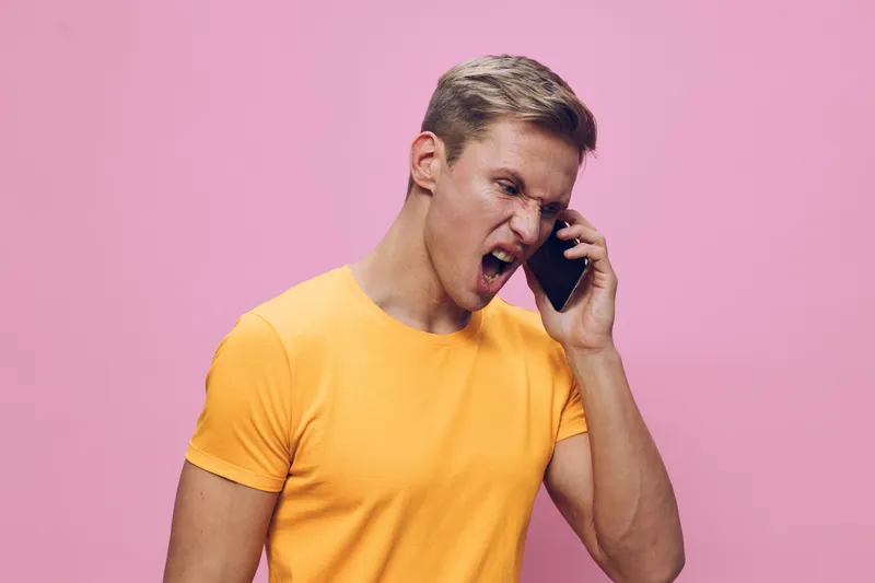 Frustrated young man with short blond hair wearing bright yellow t-shirt talking on mobile phone against pink background. Emotional facial expression, casual style, studio shot, vibrant colors, human 