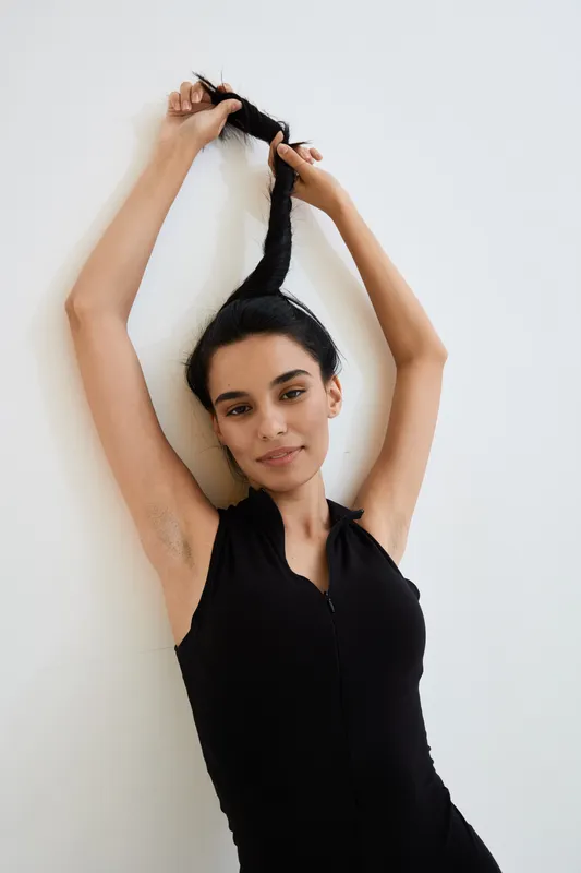 Young woman in black sleeveless top holding her long braided hair up against white background, displaying relaxed and playful mood in bright, minimal setting.