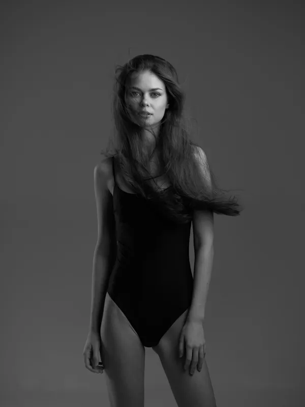 Black and white portrait of a slender woman in a black bodysuit with flowing hair against a dark background.