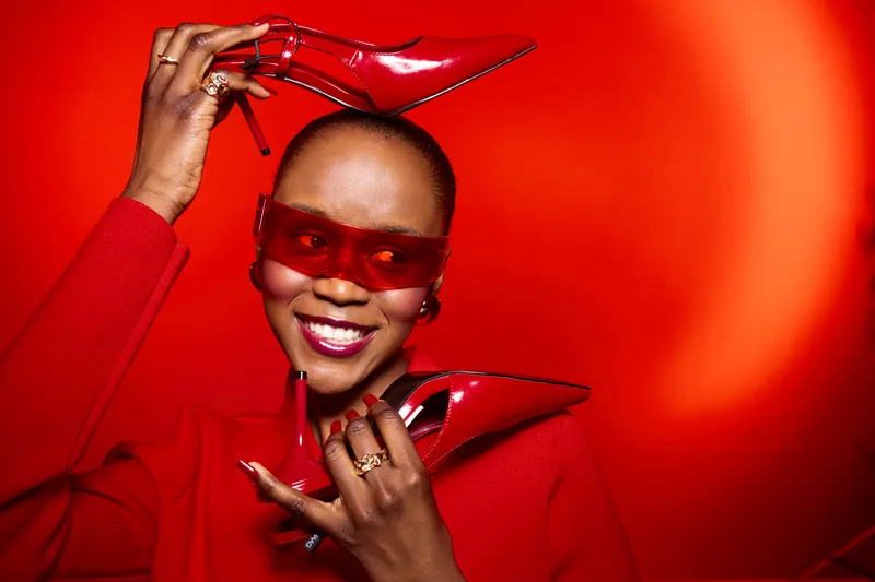 Smiling black woman with dark skin wearing red sunglasses and red outfit holds red high heels near face on red background. Concept of fashion, style, confidence, and vibrant color contrast.