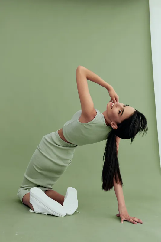Young woman posing in green knit outfit with white sneakers against soft green background, showing elegant posture and modern fashion style in studio setting.