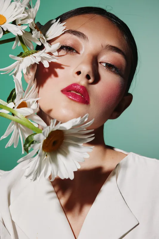 Close-up of a woman with bright makeup posing with white daisies against a mint background. Concept for beauty, fashion, creative styling, and floral elegance highlights facial glow.