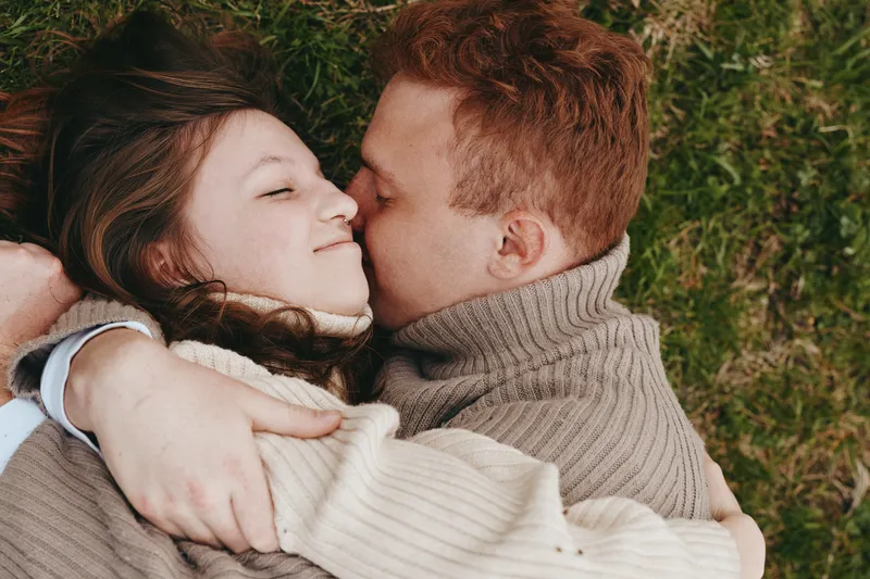 A young couple embraces closely on grass with eyes closed, sharing a tender moment of affection and warmth in cozy knit sweaters during a relaxed lifestyle scene.