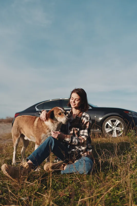 Woman sits on grassy field with a friendly dog, enjoying outdoor moments beside a sleek car, during clear sky afternoon, relaxed mood and gentle companionship in nature.