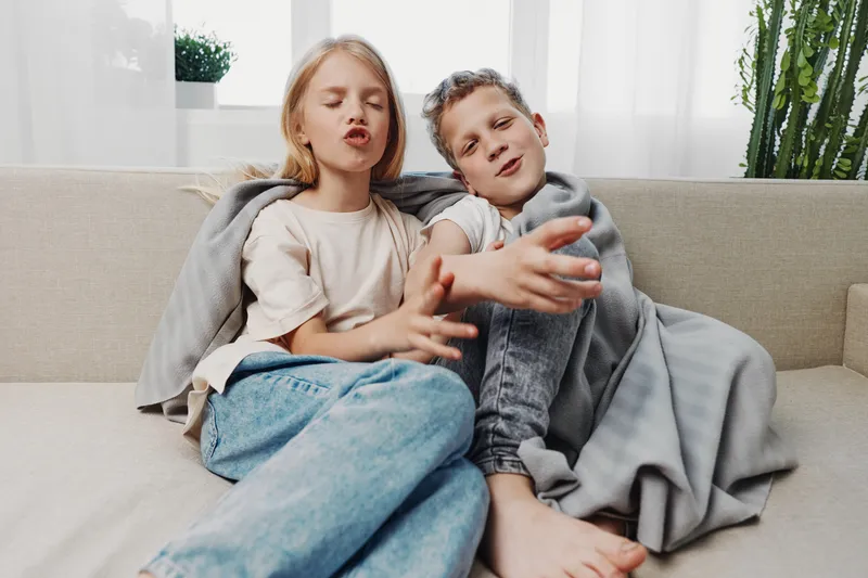 Two children sitting comfortably on a sofa covered with a blanket, playfully gesturing and enjoying cozy time indoors. Bright natural light fills the calm living room space.
