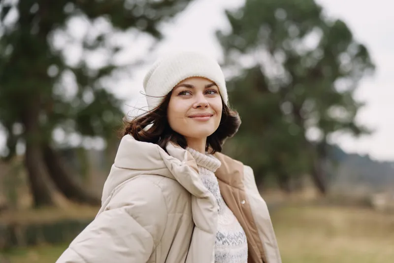 Woman in a cozy beige coat and knit beanie outdoors, smiling softly in a park setting. Casual autumn fashion portrait with natural light and calm atmosphere.