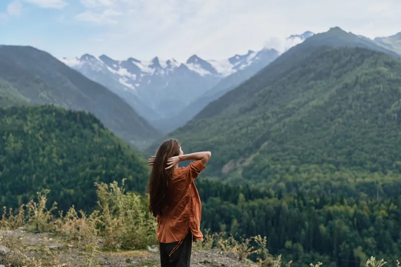 A lone traveler stands on a hillside, gazing at snow capped mountains and a vast valley, soaking in the tranquil alpine landscape and dense forest, creating a moment of awe and quiet reflection.