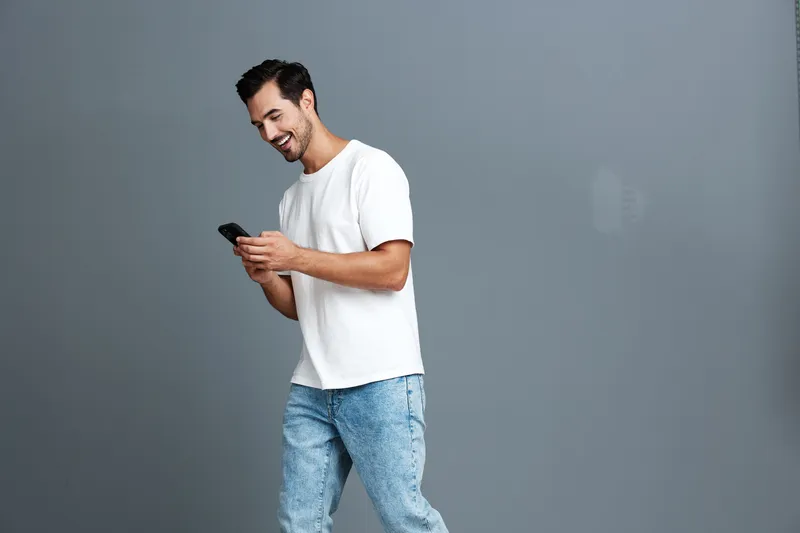 Full body smiling young man wearing white t-shirt and blue jeans, looking at his mobile phone with a cheerful expression against a plain grey background. People lifestyle concept.