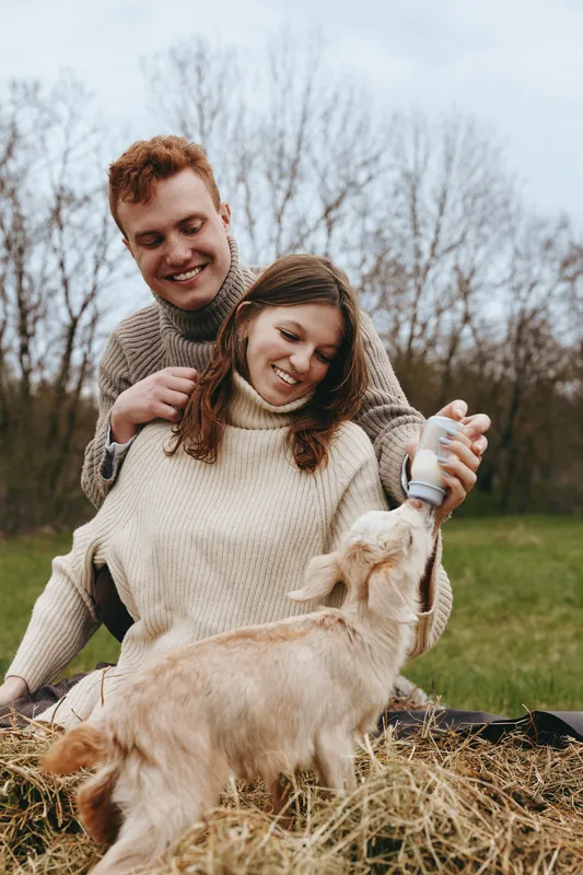 A joyful couple feeds a baby goat in a sunlit field, smiling as the kid drinks from a bottle. Casual sweaters, relaxed lifestyle, outdoor bonding, and farmyard affection.
