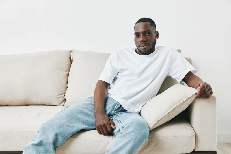 A relaxed African American man in casual attire sitting on a cozy sofa, exuding confidence and comfort, suitable for lifestyle and contemporary themes