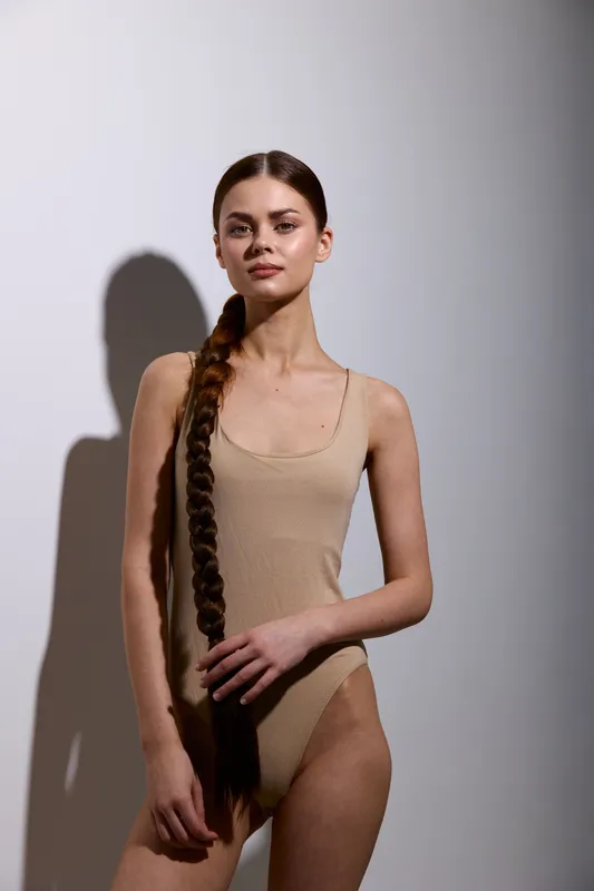 beauty woman with long braided hair wearing beige bodysuit stands against plain background with soft shadow, showcasing minimalist fashion and natural expression.