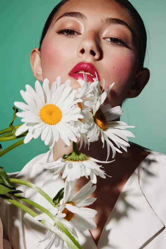 Close-up portrait of a woman with glossy red lips holding white daisies in front of her face. Soft makeup and green background. Beauty, fashion, creative concept.