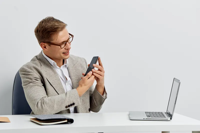 Professional young man in glasses using smartphone at modern work desk, with laptop and notebook, indoor office background, business casual attire. Focus on technology and communication concepts in a 