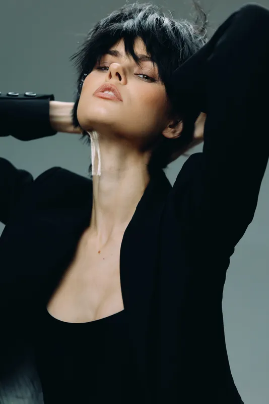 Stylish woman with short black hair posing confidently in a modern black outfit against a neutral background.