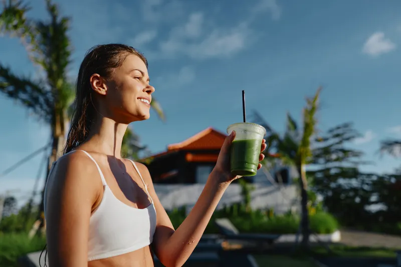 Cheerful woman outdoor smoothie drink under sunshine, summer mood, casual lifestyle moment with resort backdrop and palm trees