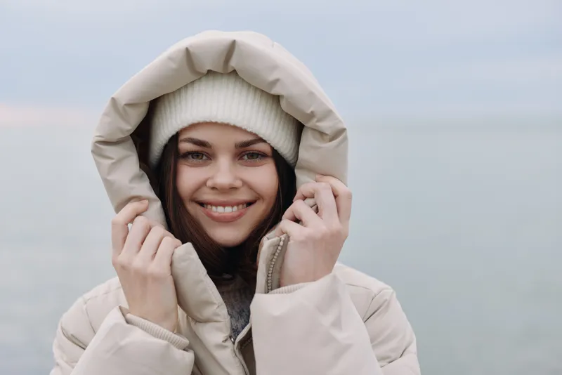 Winter woman in a hooded coat by the sea, smiling portrait with calm water and pale sky, portraying cozy outdoor chill and confident mood in cold weather with soft light and relaxed pose.