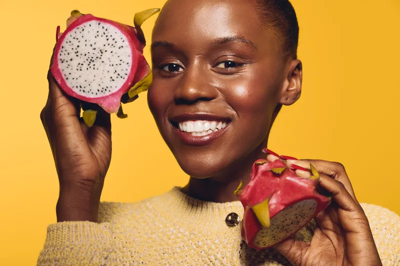 Happy African woman with dark skin holding fresh dragon fruit in both hands smiling against yellow background. Concept of healthy lifestyle, tropical fruit, and natural beauty.