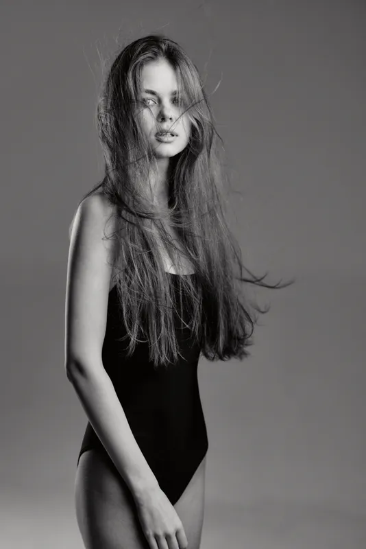 Black and white portrait of a tall woman with long hair wearing a black bodysuit and a veil, expressing calm and elegance.