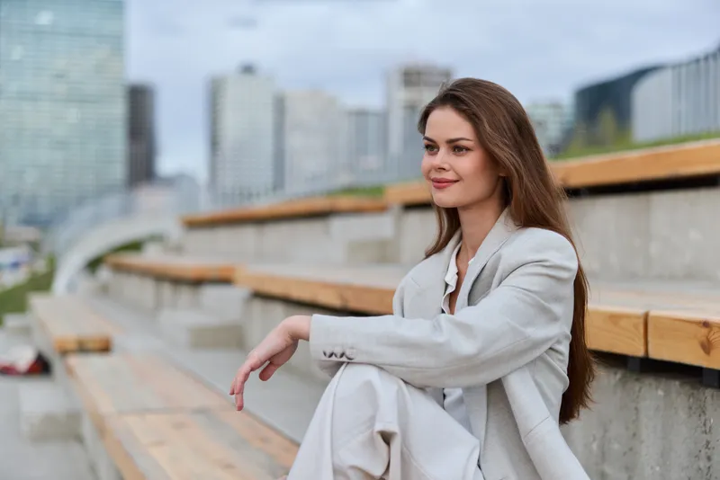 Woman in beige business suit sitting outdoors on wooden bleachers with city skyline background in soft daylight, calm confident expression