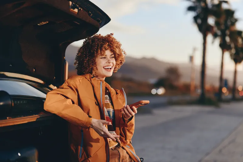 Smiling woman lifestyle shot beside a car with open door, rust jacket and curly hair, standing on a street at sunset, travel commute concept, young adult portrait, warm outdoor mood