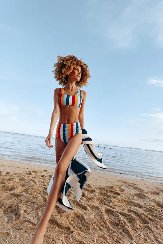 Young african american woman with curly hair walks on the sandy beach wearing a colorful striped swimsuit and holding a striped towel under a clear blue sky.