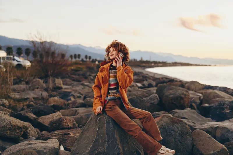 Woman lifestyle portrait at sunset on a rocky beach, curly hair, orange jacket, standing on stones with ocean horizon, warm evening light, calm leisure travel moment, relaxed smile, calm