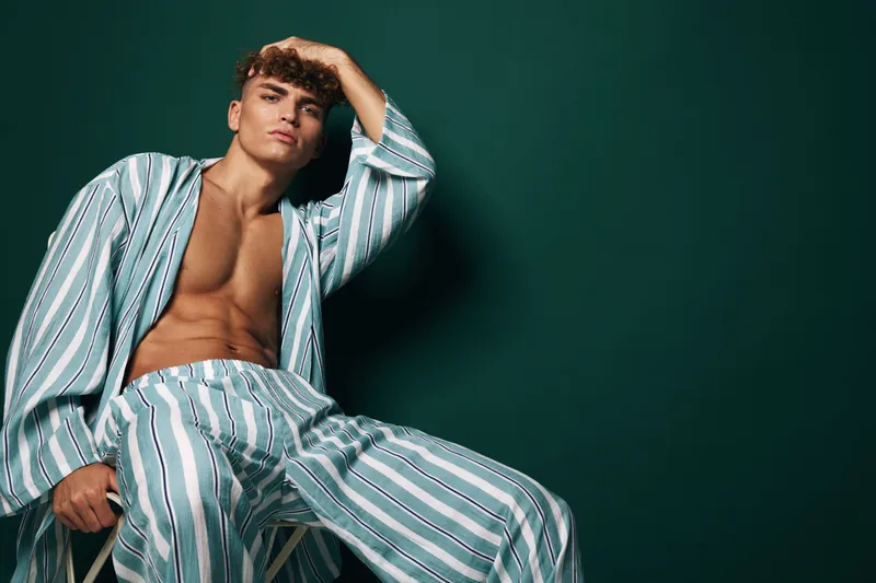 Young man in striped pajamas sitting on chair with hand running through hair against dark green wall. Relaxed male model with casual sleepwear and fresh skincare at home indoors.