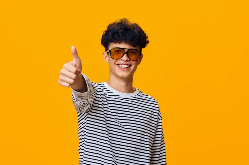 smiling man wearing striped shirt and sunglasses gives thumbs up gesture against bright yellow background, cheerful and positive expression, casual style