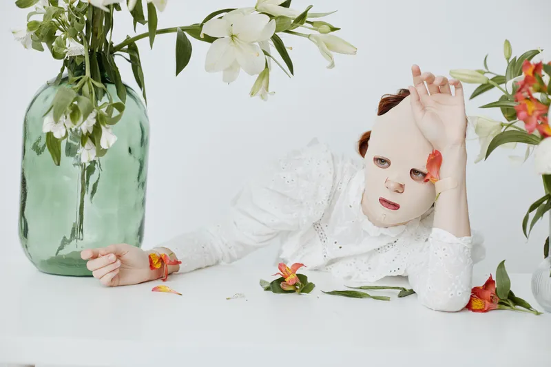 Woman in a facial mask surrounded by flowers, embodying self care and beauty in a serene, minimalistic setting with a soft color palette