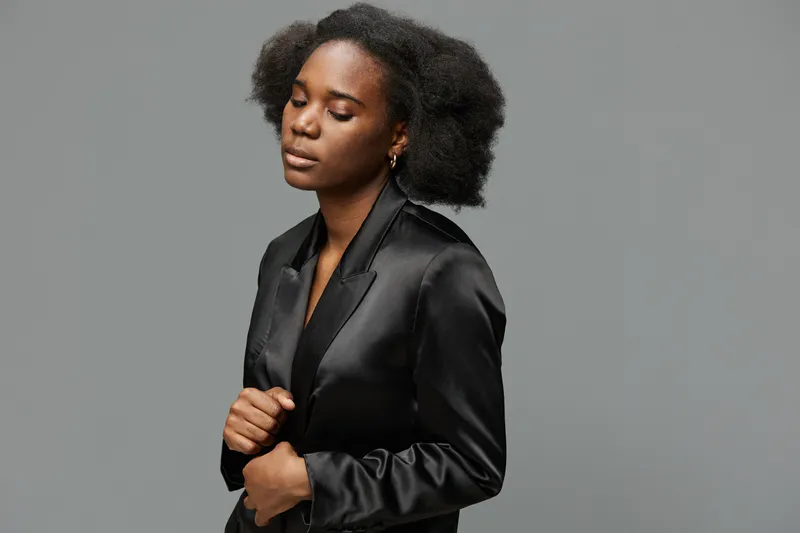 A graceful young Black woman in a sleek black satin blazer, exuding confidence and elegance, with a soft expression against a muted gray background