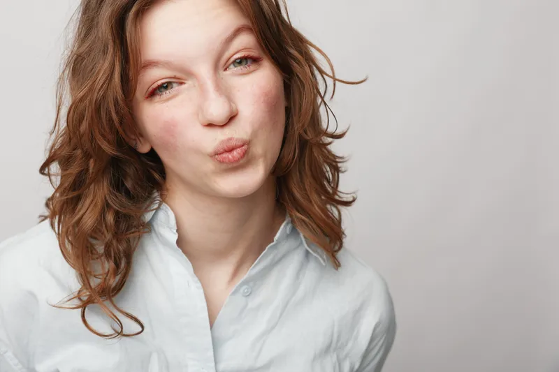 Young woman with curly hair wearing white shirt makes playful kiss face on light background. Concept of fun, youth, casual style, and positive emotions in portrait photography.