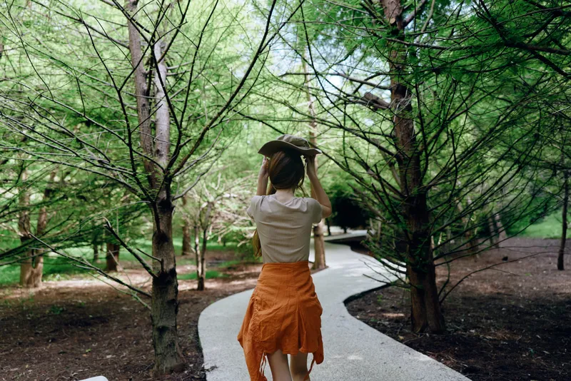 woman walking path park hat trees, back view of female in orange skirt and white top on winding paved walkway in green forest, summer stroll and casual outdoor lifestyle