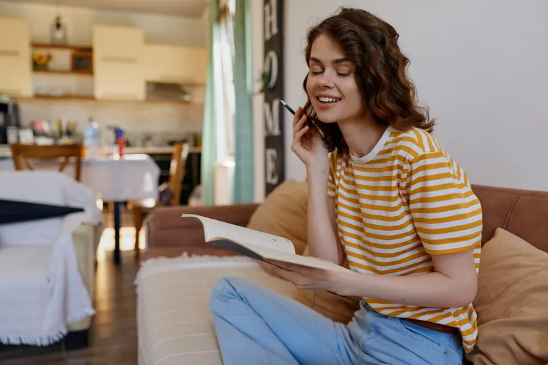 Casual young woman sits on a sofa in a cozy home living room, smiling while talking on the phone and reading a paper, enjoying a relaxed everyday moment.